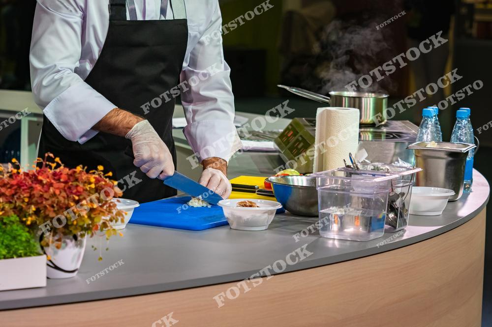 Повар в белом фартуке и перчатках нарезает овощи на кухонной станции, окруженной различными приборами, бутылками с водой и ингредиентами для приготовления еды. Атмосфера профессиональной кухни подчеркивается наличием пара и освещением.